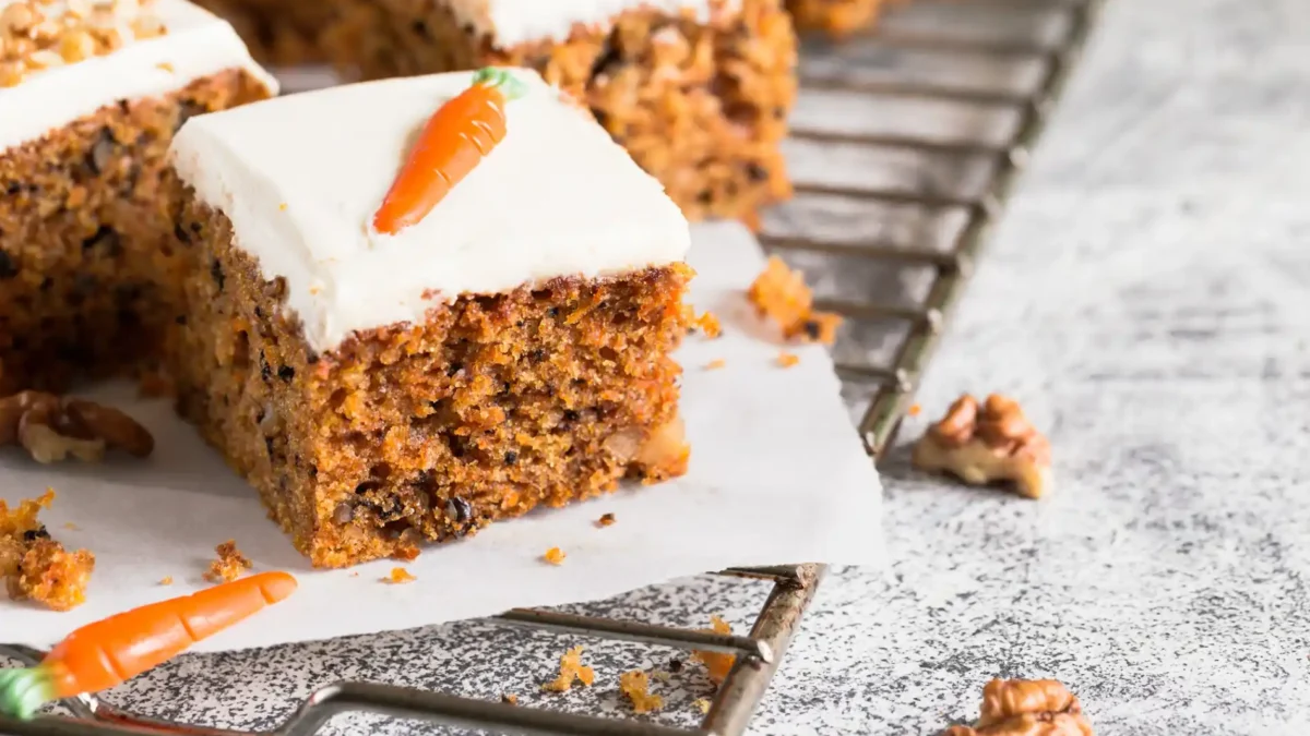 Ein detailliertes Close-up zeigt mehrere Stücke Karottenkuchen auf einem Drahtgestell, das auf einer hellen, gemusterten Oberfläche steht. Die Kuchenstücke sind mit einer dicken Schicht weißer Frischkäse-Creme überzogen und mit einer kleinen, orangefarbenen Marzipan-Karotte und gehackten Walnüssen garniert. Im Vordergrund liegt eine lose Walnusshälfte und eine Marzipan-Karotte. Die Textur des Kuchens mit den sichtbaren Karottenraspeln und Nussstücken ist gut erkennbar und wirkt appetitlich.
