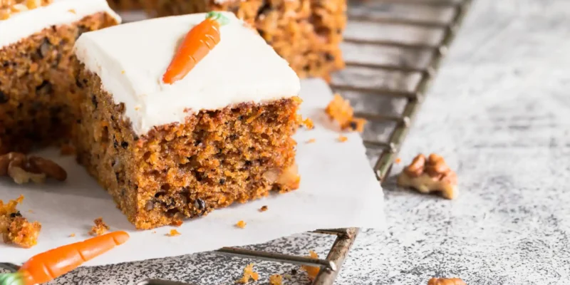 Ein detailliertes Close-up zeigt mehrere Stücke Karottenkuchen auf einem Drahtgestell, das auf einer hellen, gemusterten Oberfläche steht. Die Kuchenstücke sind mit einer dicken Schicht weißer Frischkäse-Creme überzogen und mit einer kleinen, orangefarbenen Marzipan-Karotte und gehackten Walnüssen garniert. Im Vordergrund liegt eine lose Walnusshälfte und eine Marzipan-Karotte. Die Textur des Kuchens mit den sichtbaren Karottenraspeln und Nussstücken ist gut erkennbar und wirkt appetitlich.