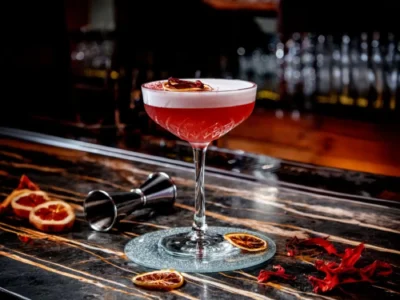Ein tiefroter Cocktail mit einer dicken, weißen Schaumkrone, garniert mit einer getrockneten Orangenscheibe und roten Blütenblättern, steht in einem Coupé-Glas auf einer eleganten, marmorierten Theke. Das Glas wird von einem glitzernden Untersetzer getragen. Links daneben liegen angeschnittene, rote Orangen und ein metallener Messbecher. Im Hintergrund verschwimmen weitere Gläser und Flaschen einer Bar. Obwohl das Getränk nicht dem klassischen Manhattan-Rezept entspricht, repräsentiert es einen eleganten und ansprechenden Bar-Klassiker wie den Manhattan Cocktail.