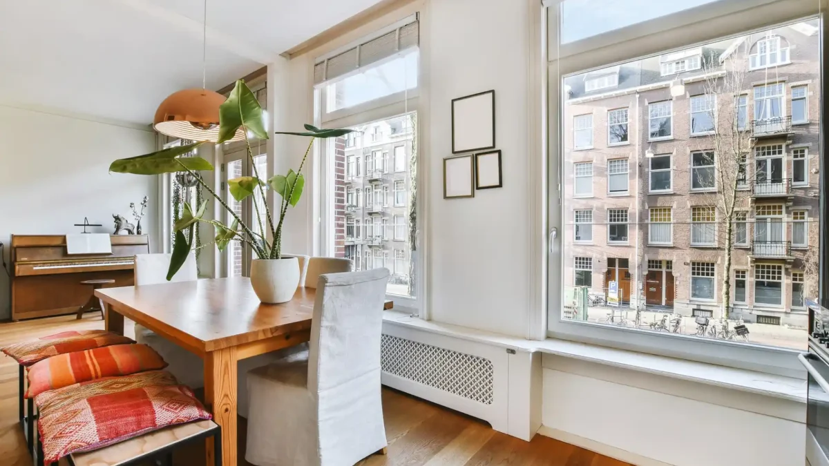 Ein lichtdurchflutetes, offenes Esszimmer zeigt eine helle Manhattan Homestyle-Einrichtung. Im Vordergrund steht ein großer Holztisch mit orange-gemusterten Sitzkissen auf einer Holzbank und einem Stuhl mit weißem Bezug. Eine große, grüne Zimmerpflanze in einem weißen Topf und eine kupferfarbene Hängelampe runden die Szene ab. Die großen Fenster geben den Blick auf eine dicht bebaute Stadtstraße frei, wodurch das urbane Flair des Stils hervorgehoben wird. Im Hintergrund ist ein Piano zu sehen, das dem Raum eine gemütliche Atmosphäre verleiht.