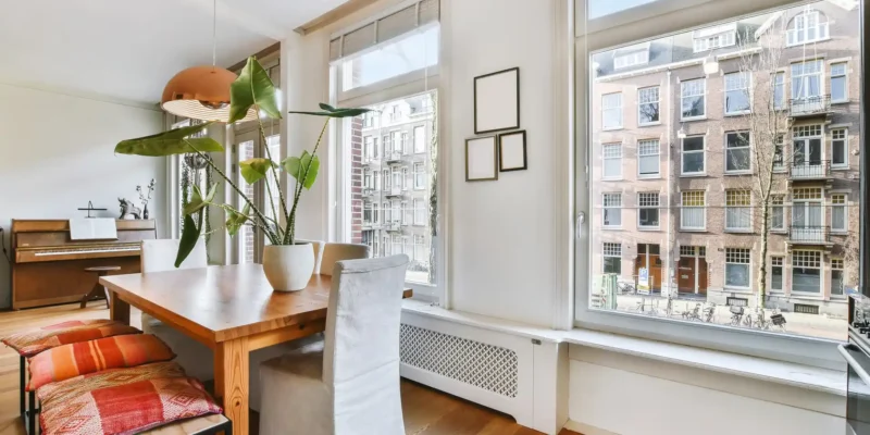 Ein lichtdurchflutetes, offenes Esszimmer zeigt eine helle Manhattan Homestyle-Einrichtung. Im Vordergrund steht ein großer Holztisch mit orange-gemusterten Sitzkissen auf einer Holzbank und einem Stuhl mit weißem Bezug. Eine große, grüne Zimmerpflanze in einem weißen Topf und eine kupferfarbene Hängelampe runden die Szene ab. Die großen Fenster geben den Blick auf eine dicht bebaute Stadtstraße frei, wodurch das urbane Flair des Stils hervorgehoben wird. Im Hintergrund ist ein Piano zu sehen, das dem Raum eine gemütliche Atmosphäre verleiht.