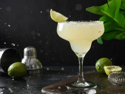 Ein appetitlicher Margarita-Cocktail in einem klassischen, breiten Margaritaglas steht auf einem braunen Holzbrett, das auf einer dunklen, nassen Oberfläche liegt. Das Glas ist am oberen Rand mit Salz bestreut und mit einer Limettenspalte garniert. Im Hintergrund stehen ein silberner Shaker und ein Sieb, neben denen ein paar Limetten und grüne Blätter liegen. Wassertropfen und Spritzer sind in der Luft und auf der Oberfläche zu sehen, was eine dynamische und erfrischende Stimmung erzeugt.