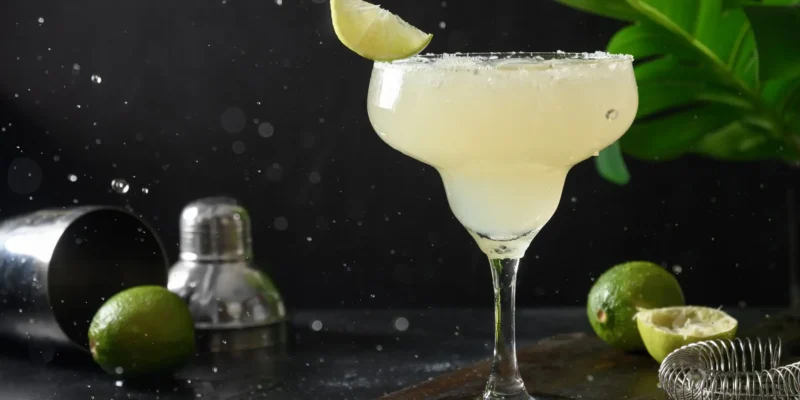 Ein appetitlicher Margarita-Cocktail in einem klassischen, breiten Margaritaglas steht auf einem braunen Holzbrett, das auf einer dunklen, nassen Oberfläche liegt. Das Glas ist am oberen Rand mit Salz bestreut und mit einer Limettenspalte garniert. Im Hintergrund stehen ein silberner Shaker und ein Sieb, neben denen ein paar Limetten und grüne Blätter liegen. Wassertropfen und Spritzer sind in der Luft und auf der Oberfläche zu sehen, was eine dynamische und erfrischende Stimmung erzeugt.