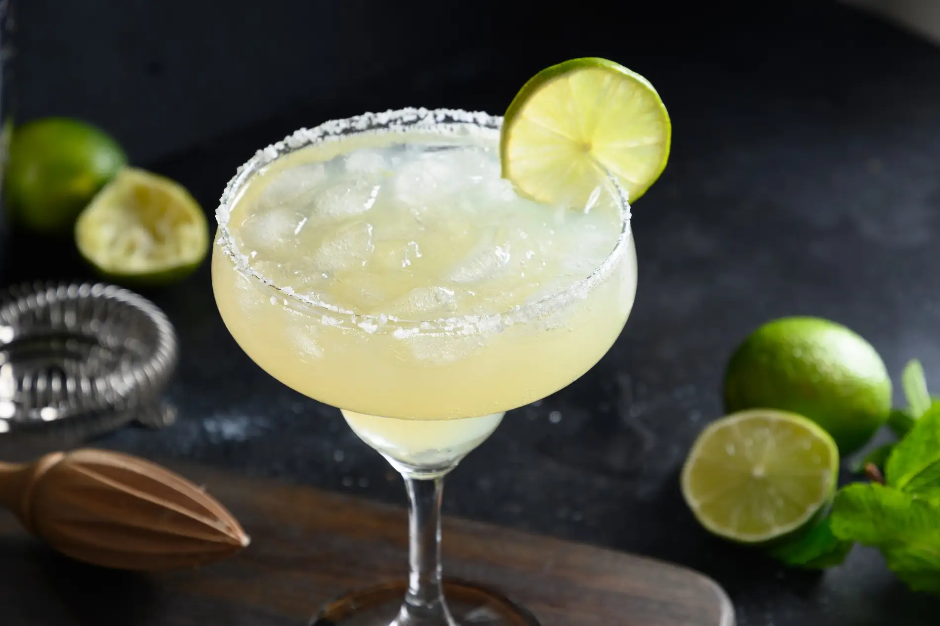 Ein klassischer, hellgelber Margarita-Cocktail in einem breiten Margaritaglas steht auf einem braunen Holzbrett, das auf einer dunklen Oberfläche mit einem leichten Salzrand liegt. Das Glas ist mit Eiswürfeln gefüllt und mit einer frischen Limettenspalte garniert. Im Hintergrund, leicht unscharf, liegen eine Handpresse für Zitrusfrüchte, eine Bar-Spirale und einige ganze sowie angeschnittene Limetten. Die Szene ist von oben aufgenommen und fängt die erfrischende und professionell zubereitete Natur des Cocktails ein.
