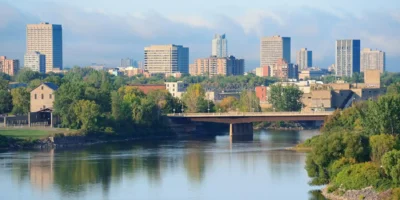 Ein Stadtpanorama zeigt hohe Gebäude und eine Brücke, die über einen breiten Fluss führt. Die Bäume im Vordergrund und an den Ufern sind in kräftigen Grüntönen gehalten. Der Himmel ist klar und blau mit einigen Wolken. Obwohl es sich nicht um eine Stadt in Pennsylvania handelt (sondern wahrscheinlich um Winnipeg, Manitoba), visualisiert das Bild eine typische urbane Flusslandschaft, wie sie in den Städten Pennsylvanias zu finden ist.