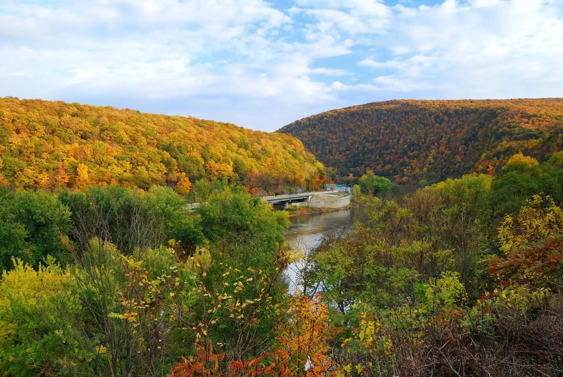 Ein weitläufiges Panorama zeigt eine malerische Herbstlandschaft, die typisch für Pennsylvania sein könnte. Ein breiter Fluss fließt durch ein Tal, das von bewaldeten Hügeln flankiert wird, deren Bäume in kräftigen Gelb-, Orange- und Rottönen leuchten. Eine Brücke führt über den Fluss und verbindet die beiden Ufer. Der Himmel darüber ist hellblau mit einigen weißen Wolken. Das Bild fängt die Schönheit und die herbstliche Atmosphäre der Natur in der Region ein.