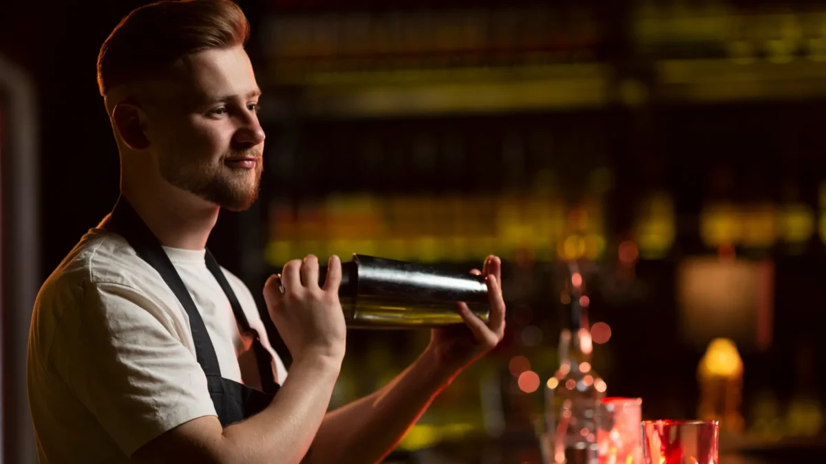 Ein gut beleuchteter Barkeeper mit gepflegtem Bart und einem weißen T-Shirt unter einer dunklen Schürze schüttelt konzentriert einen metallischen Cocktail-Shaker. Er steht vor einem unscharfen, dunklen Hintergrund, der eine Bar mit beleuchteten Flaschenregalen andeutet. Die Szene symbolisiert die handwerkliche Präzision des Mixens, eine Fähigkeit, die durch historische Werke wie den Bartender’s Guide von Jerry Thomas etabliert wurde.