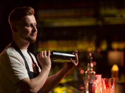 Ein gut beleuchteter Barkeeper mit gepflegtem Bart und einem weißen T-Shirt unter einer dunklen Schürze schüttelt konzentriert einen metallischen Cocktail-Shaker. Er steht vor einem unscharfen, dunklen Hintergrund, der eine Bar mit beleuchteten Flaschenregalen andeutet. Die Szene symbolisiert die handwerkliche Präzision des Mixens, eine Fähigkeit, die durch historische Werke wie den Bartender’s Guide von Jerry Thomas etabliert wurde.