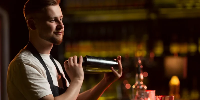 Ein gut beleuchteter Barkeeper mit gepflegtem Bart und einem weißen T-Shirt unter einer dunklen Schürze schüttelt konzentriert einen metallischen Cocktail-Shaker. Er steht vor einem unscharfen, dunklen Hintergrund, der eine Bar mit beleuchteten Flaschenregalen andeutet. Die Szene symbolisiert die handwerkliche Präzision des Mixens, eine Fähigkeit, die durch historische Werke wie den Bartender’s Guide von Jerry Thomas etabliert wurde.