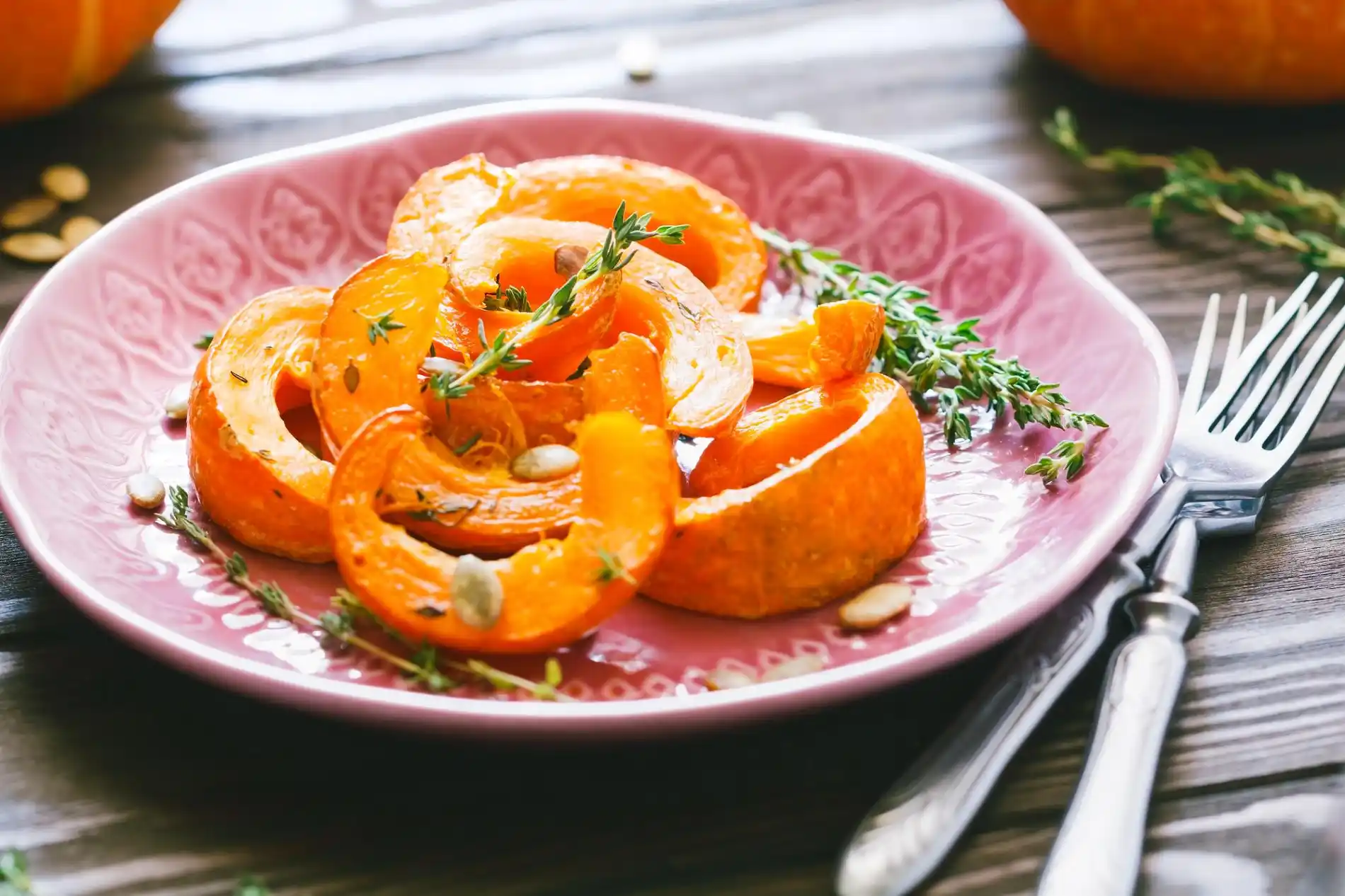 Ein rustikales Close-up zeigt auf einem rosa-gemusterten Teller ein Gericht aus goldgelben, halbmondförmigen Kürbisscheiben, die mit frischen Thymianzweigen und Kürbiskernen garniert sind. Das Gericht ist wahrscheinlich ein gerösteter oder gebackener Kürbis, der eine Beilage zu herzhaften Gerichten sein könnte. Die Szene, die Kürbis in einer eleganten und aromatischen Form präsentiert, ist eine Anregung für vielseitige Crown Prince Kürbis Rezepte. Besteck und eine Holztischoberfläche rahmen den Teller ein.