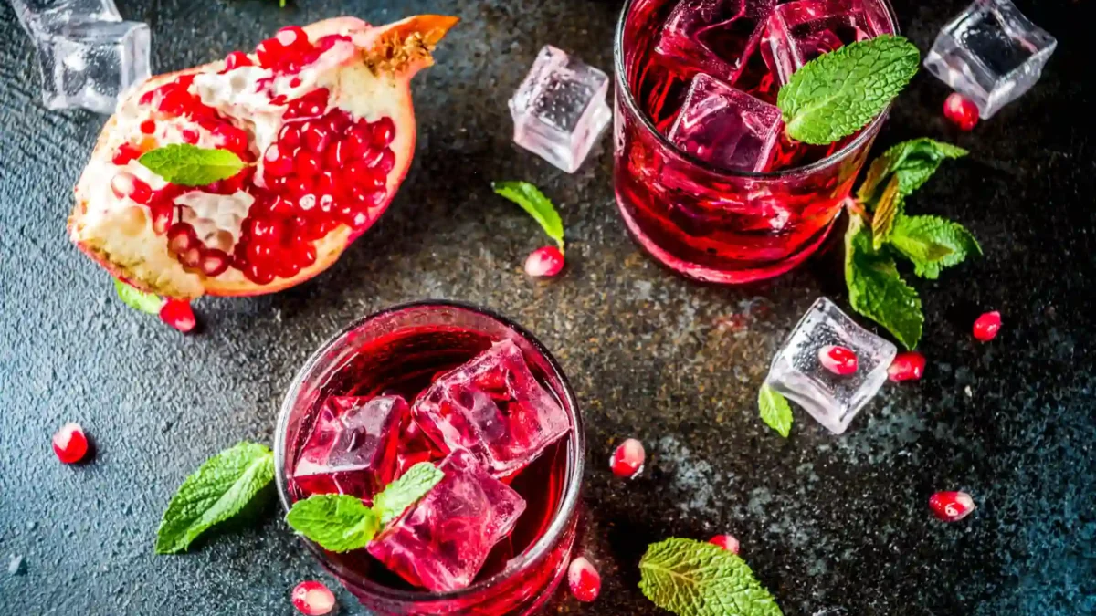 Ein dramatisches Flat-Lay-Foto zeigt zwei Tumbler-Gläser, gefüllt mit dem leuchtend rubinroten New York Sour, da er mit Rotwein zubereitet wird. Die Drinks sind mit klaren Eiswürfeln und frischen Minzblättern garniert. Daneben liegt eine halbierte, aufgeschnittene Granatapfelfrucht, deren Kerne lose um die Gläser verstreut sind. Das dunkle, strukturierte Hintergrundthema erzeugt einen starken Kontrast zur hellen Minze und den roten Granatapfelkernen, was die Komplexität und Eleganz des Cocktails unterstreicht.