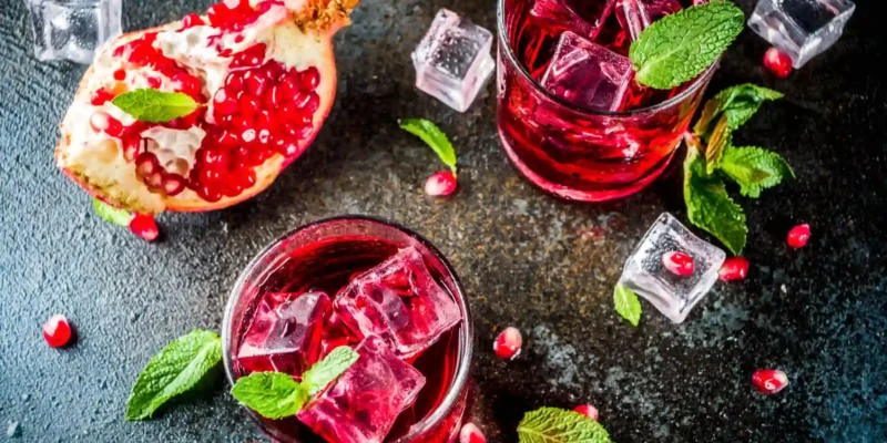 Ein dramatisches Flat-Lay-Foto zeigt zwei Tumbler-Gläser, gefüllt mit dem leuchtend rubinroten New York Sour, da er mit Rotwein zubereitet wird. Die Drinks sind mit klaren Eiswürfeln und frischen Minzblättern garniert. Daneben liegt eine halbierte, aufgeschnittene Granatapfelfrucht, deren Kerne lose um die Gläser verstreut sind. Das dunkle, strukturierte Hintergrundthema erzeugt einen starken Kontrast zur hellen Minze und den roten Granatapfelkernen, was die Komplexität und Eleganz des Cocktails unterstreicht.