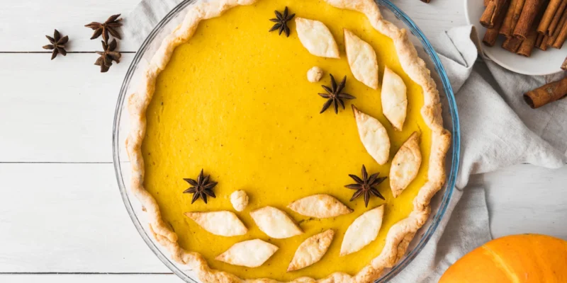 Ein perfekt runder Pumpkin Pie befindet sich in einer transparenten Glasform und ist von oben aufgenommen. Die Füllung leuchtet in einem satten, tiefen Gelb-Orange und ist dekorativ mit Sternanis und kleinen, blattförmigen Teigstücken verziert. Der Rand des Mürbeteigs ist leicht gewellt und goldbraun. Der Pie liegt auf einem hellgrauen Tuch, umgeben von Zimtstangen und weiteren Sternanis-Gewürzen. Im unteren rechten Bereich ist ein Teil eines orangefarbenen Kürbisses sichtbar. Die Szene ist hell beleuchtet und symbolisiert ein gelungenes, festliches Pumpkin Pie Rezept vegan.